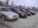2003 Ford Crown Victoria - A line of Ford Crown Victorias, including a silver model in the foreground, are parked with a city skyline in the background.