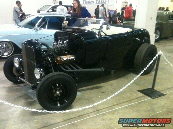 black hot rod roadster with exposed engine and wide rear tires, parked indoors with a light blue car and other people nearby