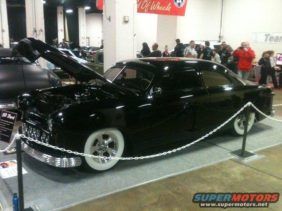 Black 1950 Ford coupe with whitewall tires and chrome grille, hood open, at a car show