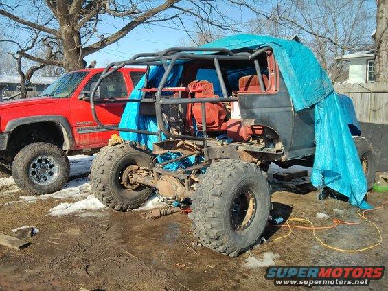 Off-road vehicle chassis with roll cage and blue tarp, next to a red pickup truck