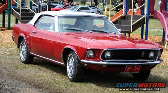 Red convertible classic car with white top parked on grass near playground equipment. License plate frame reads '70s 69'