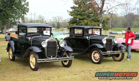 Two black vintage cars, one a pickup truck, parked on grass. The car on the right has a man standing next to it