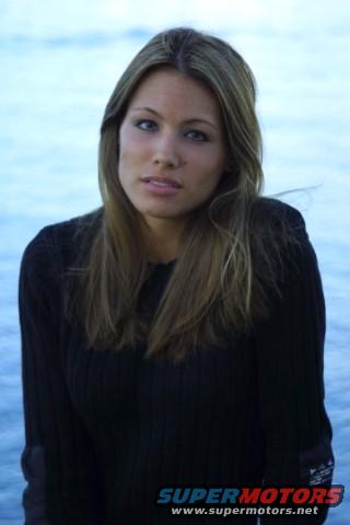 Woman with long blonde hair wearing a black sweater, standing in front of a body of water. Supermotors.net logo visible