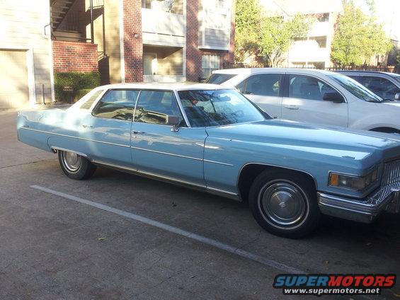 Light blue 1975 Cadillac Sedan de Ville with a white vinyl roof, parked next to a white SUV