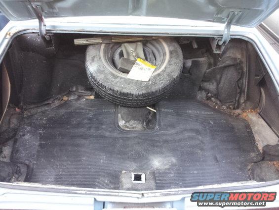 Car trunk interior with a spare tire, jack, and worn floor mat. Light blue exterior paint visible