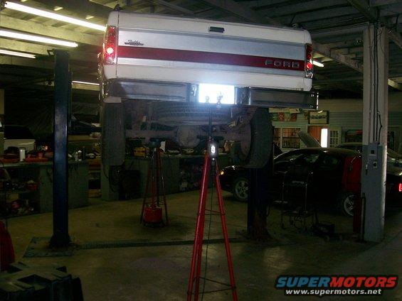Rear view of a white Ford pickup truck on a lift, with a red stripe and 'FORD' lettering. Another car is visible below