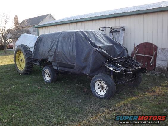 Vehicle chassis covered by a tarp, with a tractor behind it. Front end is exposed