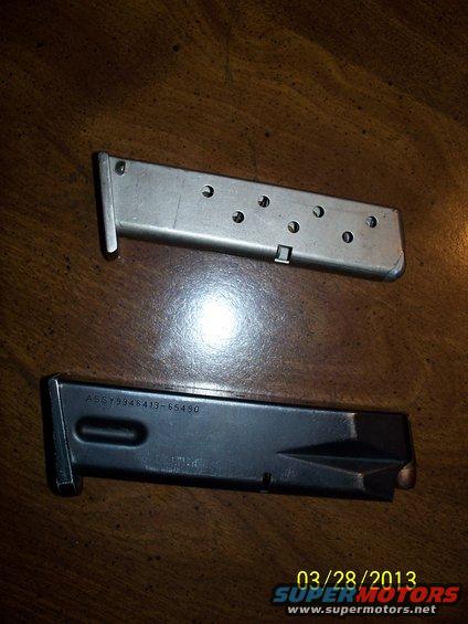 Two gun magazines, one silver and one black, on a wooden surface. The black magazine has text that reads ASSY9346413-65490