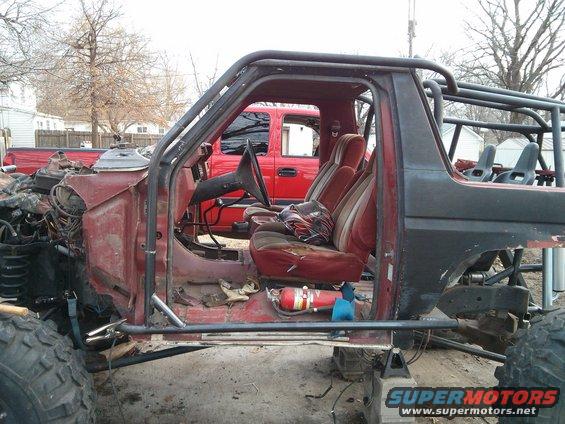 Modified SUV interior with roll cage, red seats, and exposed engine components; red pickup truck in background