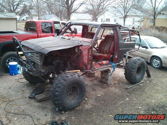 1986 Ford Bronco project vehicle with roll cage, large tires, and removed body panels. Red pickup truck in background