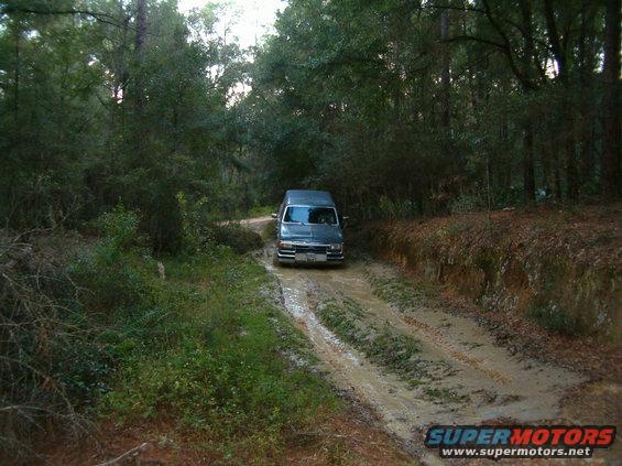 Dark colored van driving on a muddy, rutted dirt road through a wooded area