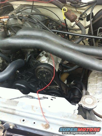 Engine bay with hoses, belts, and engine components visible. A red wire runs across the engine