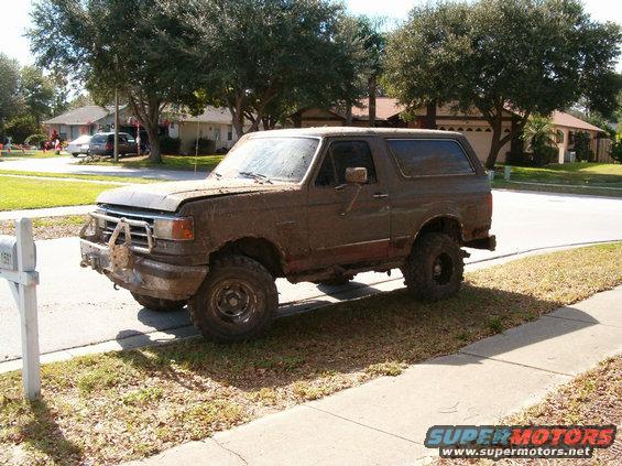 Mud-covered brown Ford Bronco with aftermarket bumper parked on grass, 3/4 front view