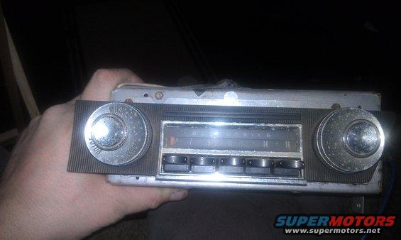 Hand holding an old car radio with two knobs and push buttons. The tuning dial shows 'OLDSMOBILE'