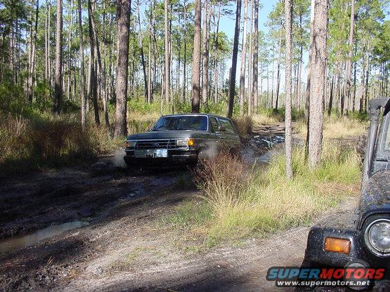 Dark green Ford Bronco driving through mud, front quarter view. Another vehicle is partially visible on the right