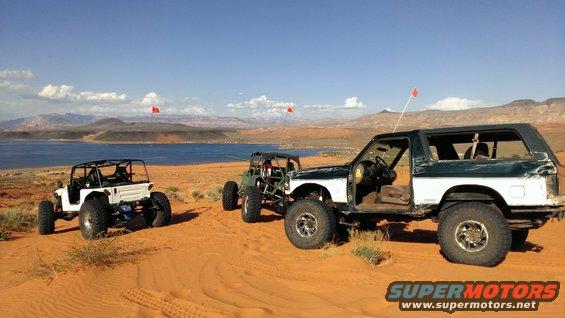 Three modified off-road vehicles parked on sand dunes with a lake and mountains in the background