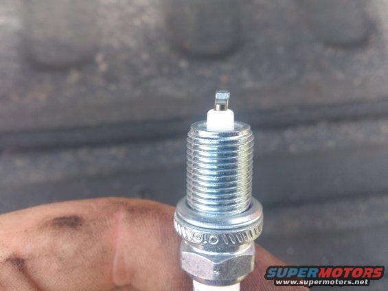 Spark plug held by a dirty hand against a blurred background. Text 'SUPERMOTORS' is visible