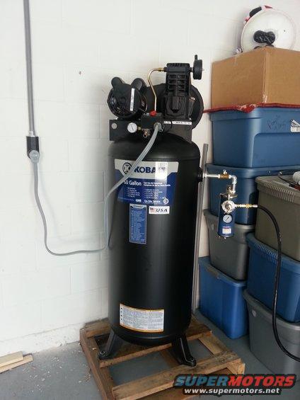 Black Koba 60 gallon air compressor on wooden pallet, next to storage bins and a cardboard box