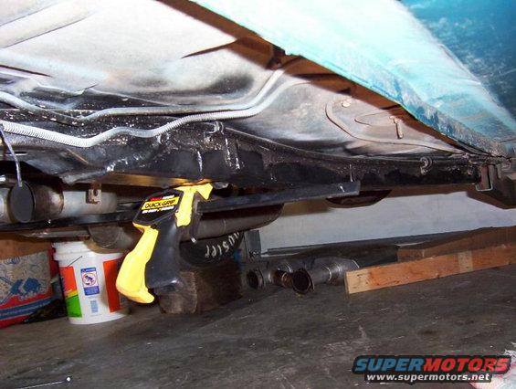under a car showing exhaust pipes, fuel tank, and a clamp holding something