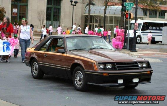 Brown 1980 Ford Mustang driving down street with people walking nearby