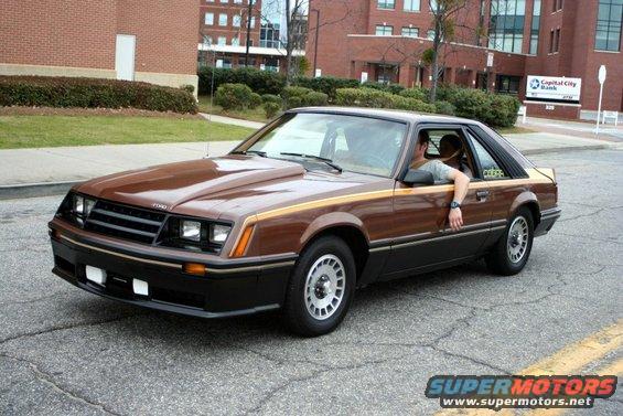 Brown 1980 Ford Mustang coupe with a hood scoop and gold side stripe, viewed from the front three-quarter angle