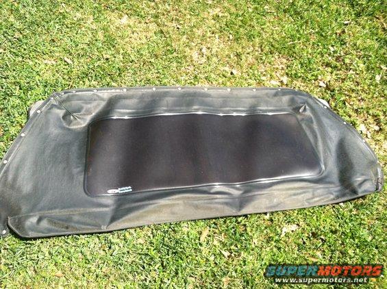 Black soft top with a clear plastic rear window, laid out on grass. Small logo visible on the window frame