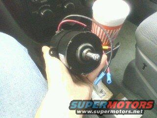 Hand holding a black blower motor with red and blue wires attached. Supermotors.net logo visible