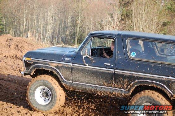 Black Ford Bronco driving through mud, tires kicking up dirt. Driver visible inside