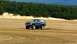 1979 Ford Bronco - A dark SUV with a lifted suspension drives across a sandy landscape with a forest in the background.
