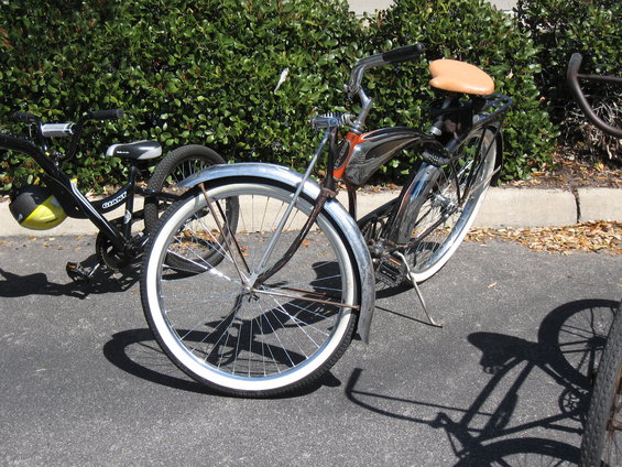 Chrome cruiser bicycle with tan seat and whitewall tires, parked next to a black bicycle with a yellow helmet
