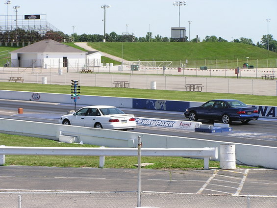 Two sedans race on a drag strip, one white and one dark blue