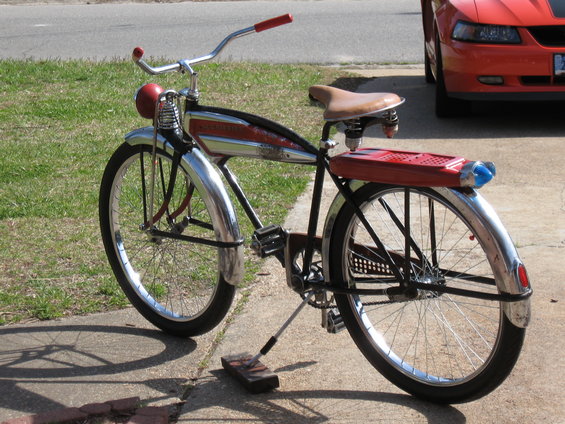 Side view of a vintage red and chrome bicycle with a red rear rack and blue taillight, parked next to an orange car
