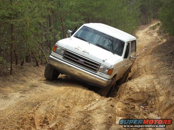 1990 Ford Bronco navigating a steep, muddy off-road trail with trees on either side