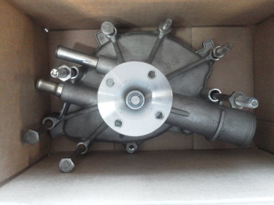 Engine water pump with silver flange and metal pipes, inside a cardboard box