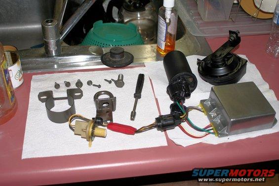Various automotive electrical parts including a black ignition coil, a silver control module, a horn, and assorted screws and brackets laid out on a paper towel