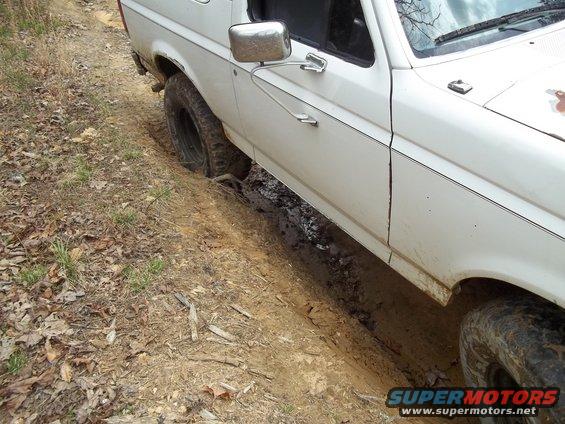 White SUV with one wheel in a muddy rut on a dirt path