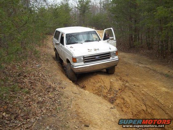 White Ford Bronco driving off-road on a dirt track, driver's door open