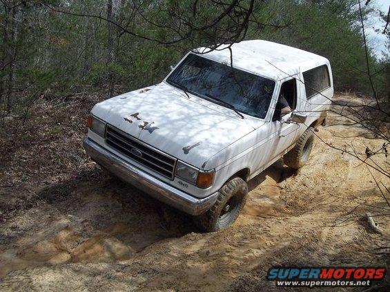White 1987 Ford Bronco on a dirt trail, front-left quarter view, with rust spots on hood