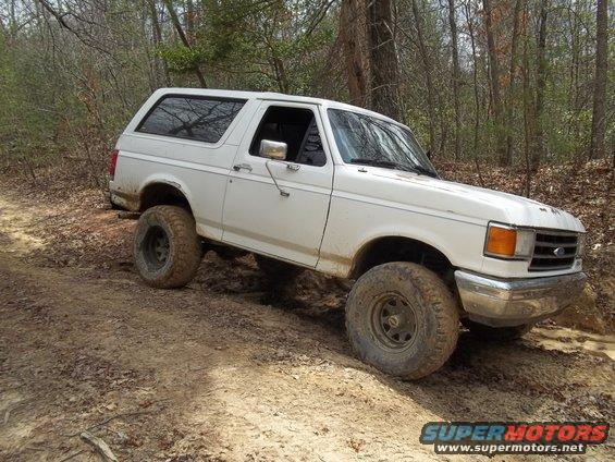 White 1990 Ford Bronco with aftermarket wheels and tires on a muddy trail