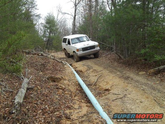 White SUV driving on a dirt path in a wooded area. The front grille and headlights are visible