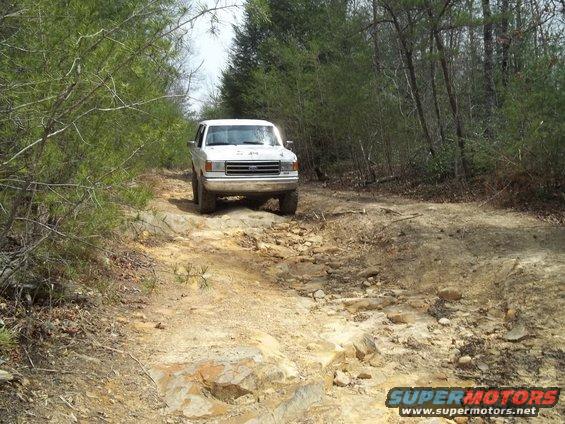 White 1990 Ford Bronco driving on a rocky, wooded trail