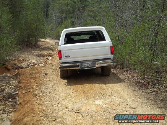 White Ford Bronco driving on a dirt road, rear view. Chrome bumper, license plate visible