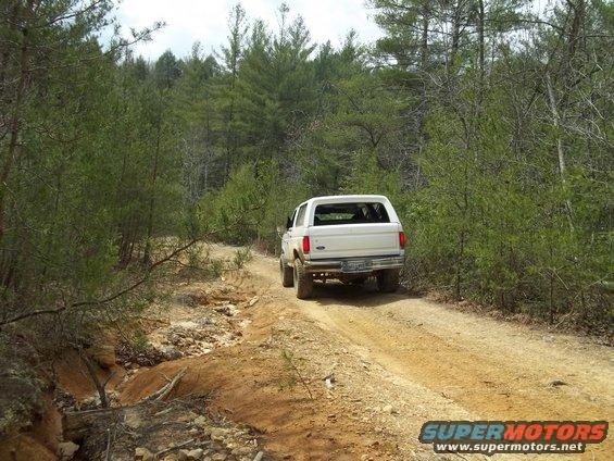 White 1990 Ford Bronco driving on a dirt road in a forest