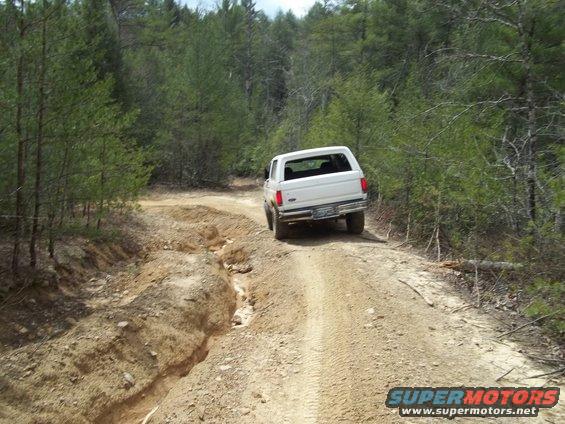 White 1990 Ford Bronco driving on a rutted dirt road through trees