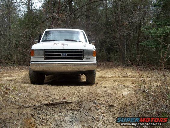 Front view of a white Ford F-series truck on a dirt road in the woods. Grille indicates 1987 model year