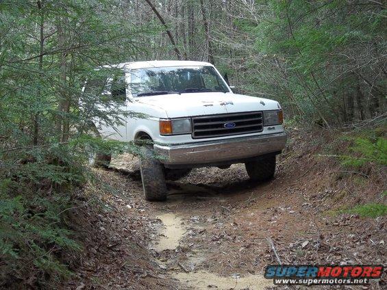 White Ford truck driving on a dirt path in the woods. Front grille and bumper are visible