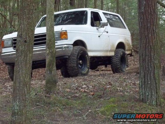 White 1990 Ford Bronco with off-road tires driving through trees