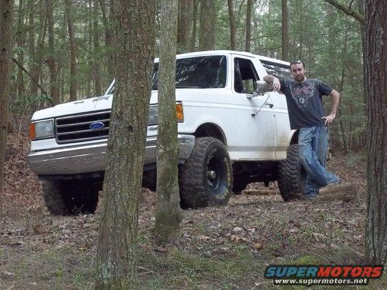 White 1990 Ford Bronco parked off-road in a wooded area with a man leaning against it