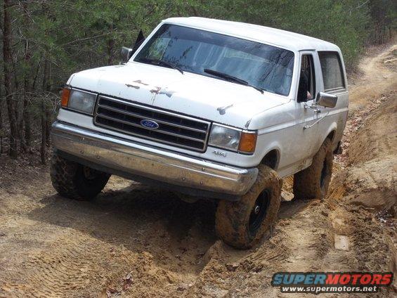 White Ford Bronco with mud-caked tires navigating a dirt trail, front-left tire high
