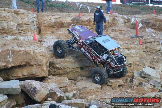Custom off-road buggy with large tires navigating a rocky incline. Multiple sponsor decals adorn the body